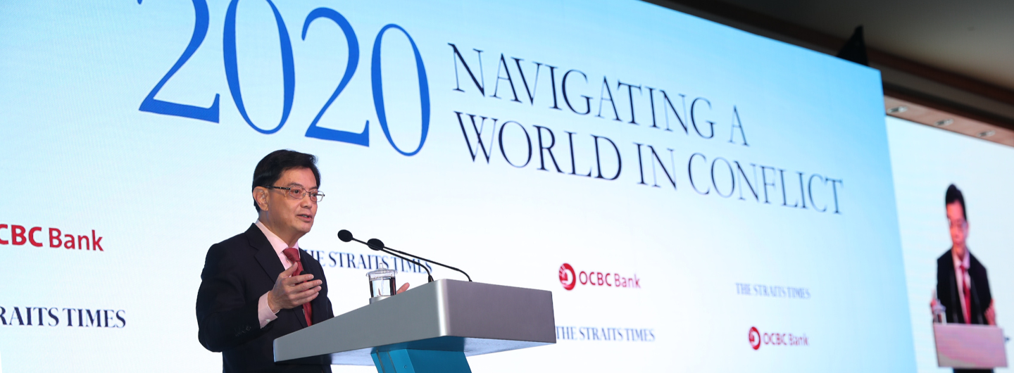 Man in suit speaking at podium; "2020 Navigating a World in Conflict" backdrop, OCBC Bank and Straits Times logos.